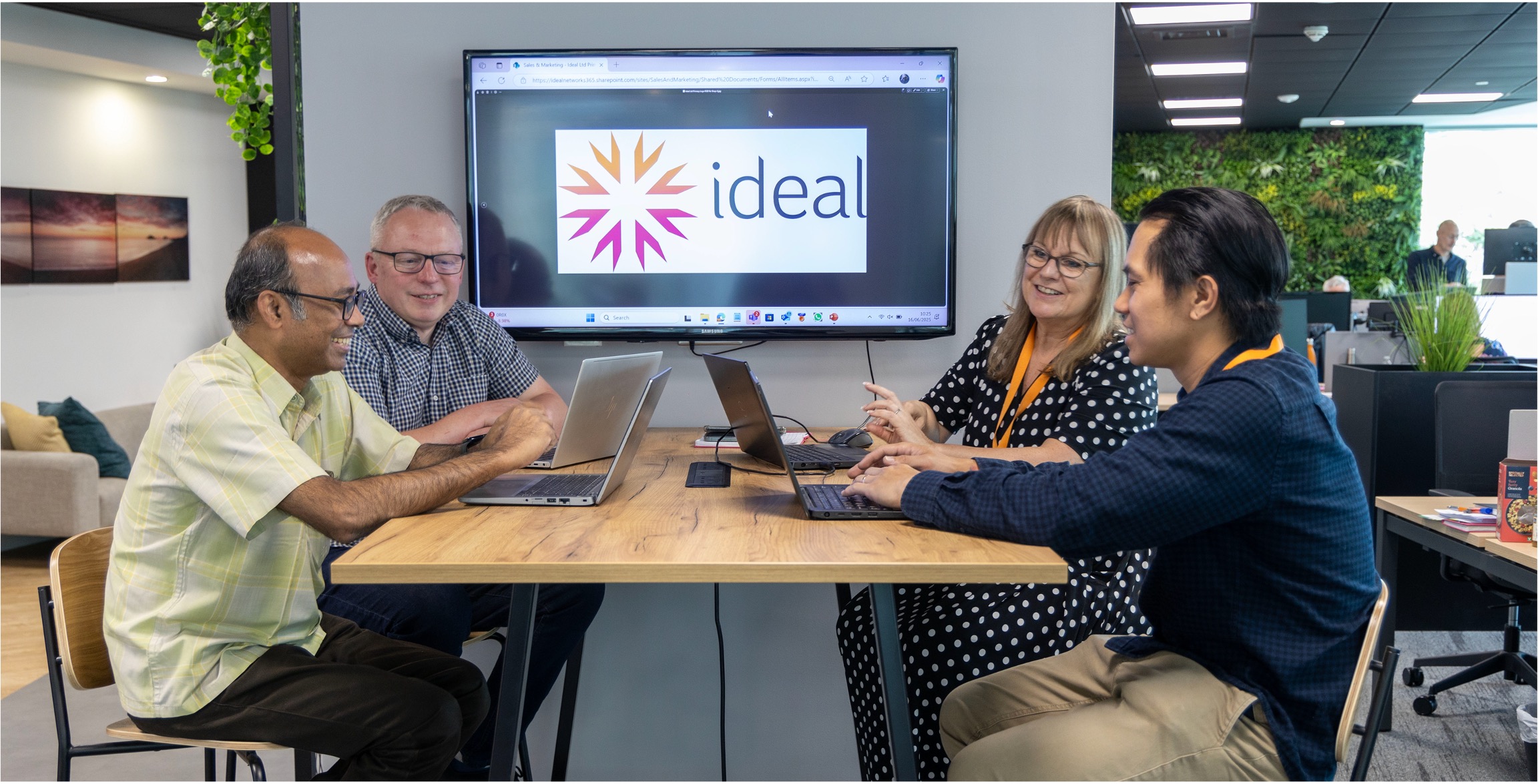 Four colleagues in the Ideal offices discuss a project at a table, with a screen behind.